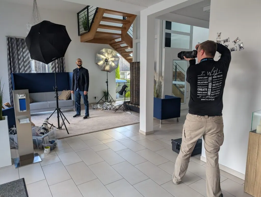 Fotograf bei Porträtaufnahmen im Büro, Schirme und Blitzlicht aufgebaut, ein Mitarbeiter steht vor dem Sofa