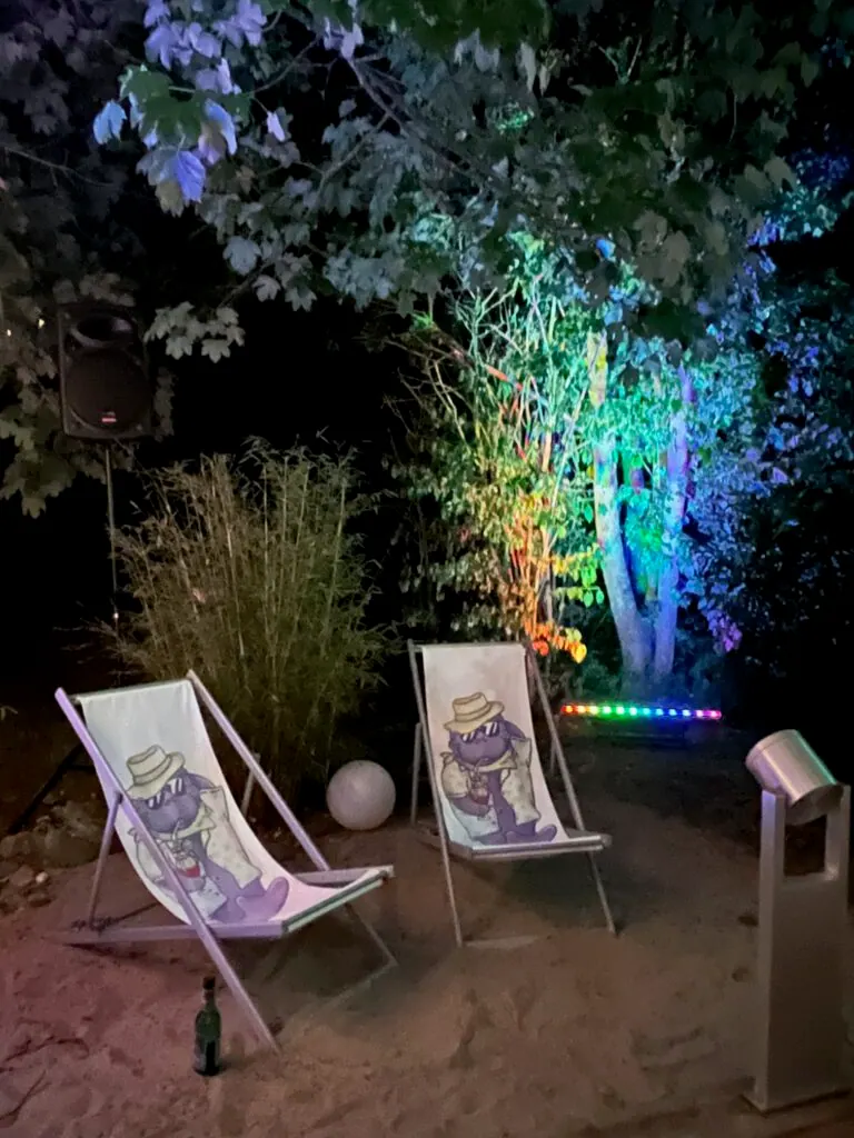 Zwei Liegestühle mit Vera-Motiv im Sand, bunt beleuchtete Bäume bei Nacht beim Sommerfest 2025