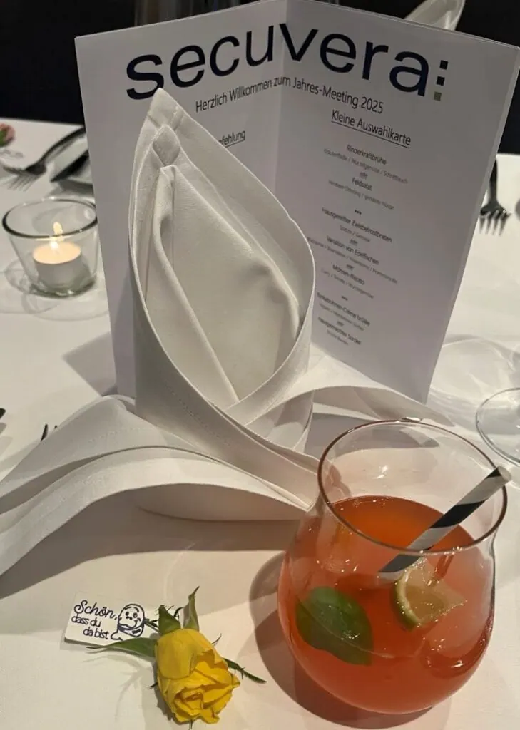Gedeckter Tisch mit secuvera-Speisekarte zum Jahres-Meeting 2025, Cocktail und gelber Rose