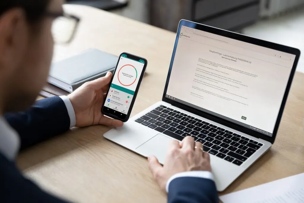 Person hält ein Smartphone mit der PANDERAM-App, auf dem Laptop läuft die Umfrage der Laborerhebung