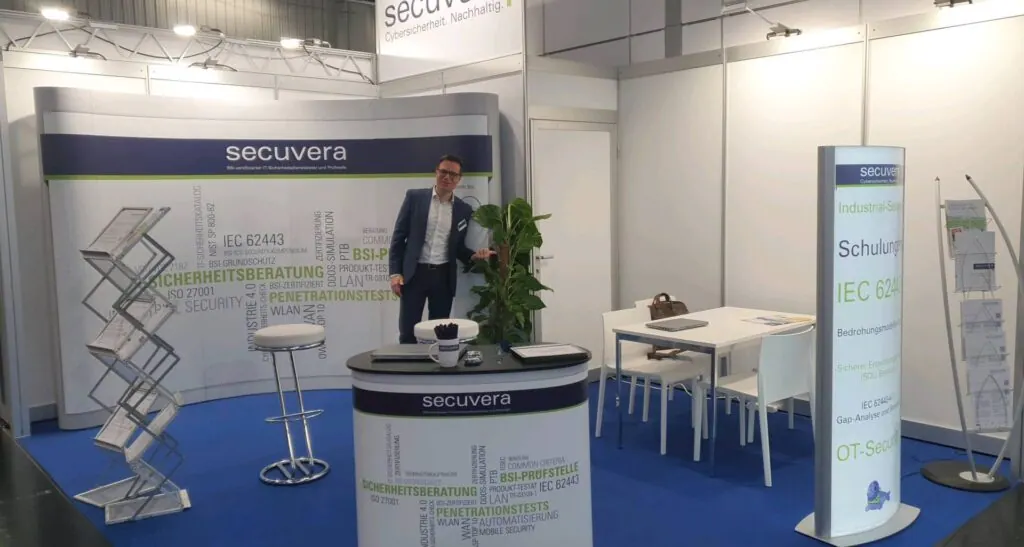 secuvera-Messestand auf der SPS 2023 mit Mitarbeiter, Infowand zu Sicherheitsberatung und Penetrationstests, Prospektständern