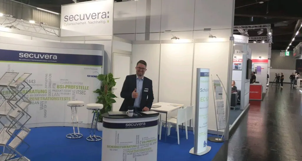 Mitarbeiter von secuvera zeigt Daumen hoch am Messestand der SPS 2023 mit Stehtisch und Infostele zu IEC 62443