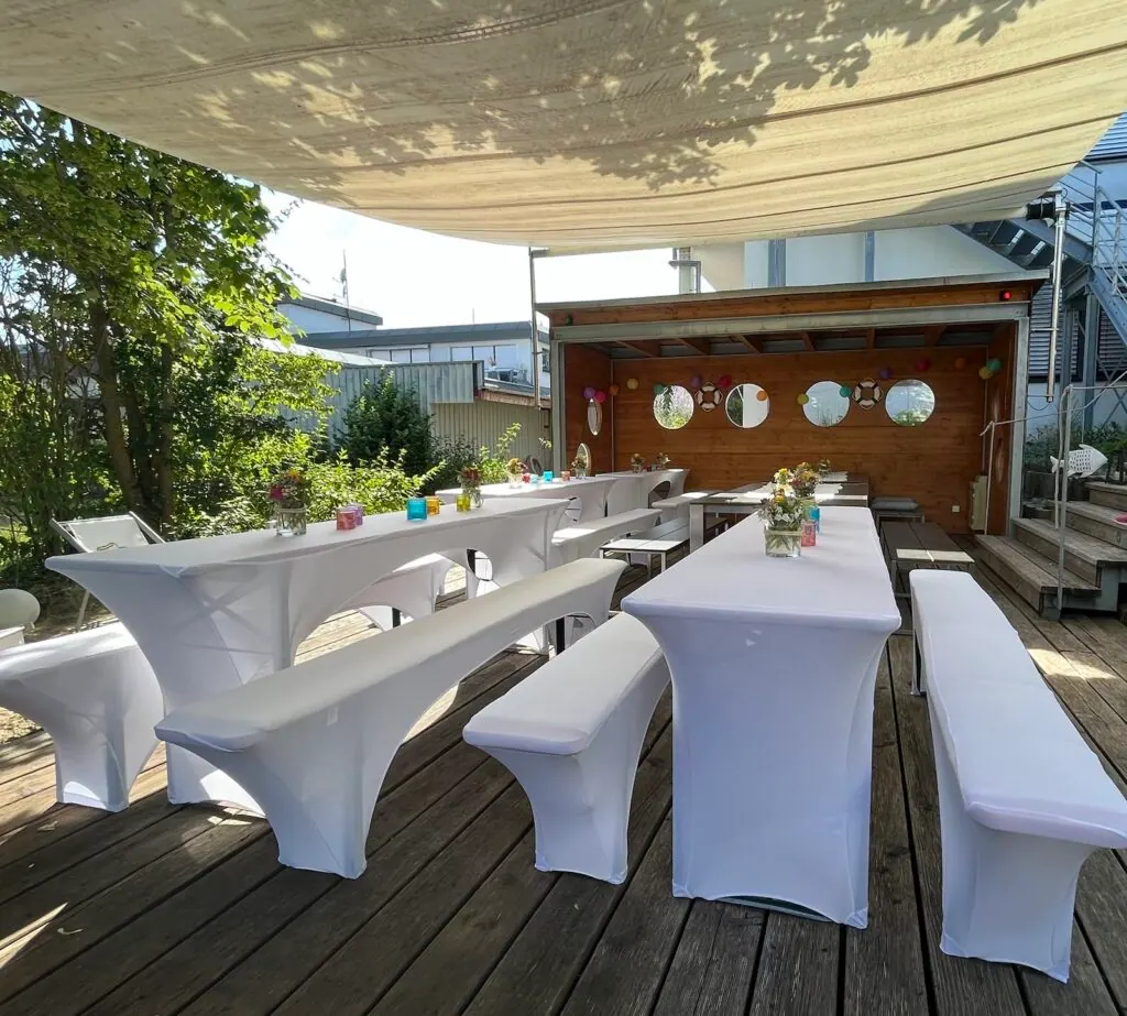 Terrasse mit weiß gedeckten Tischen und Bänken, im Hintergrund eine Holzhütte mit Bullaugen, für das Sommerfest 2023