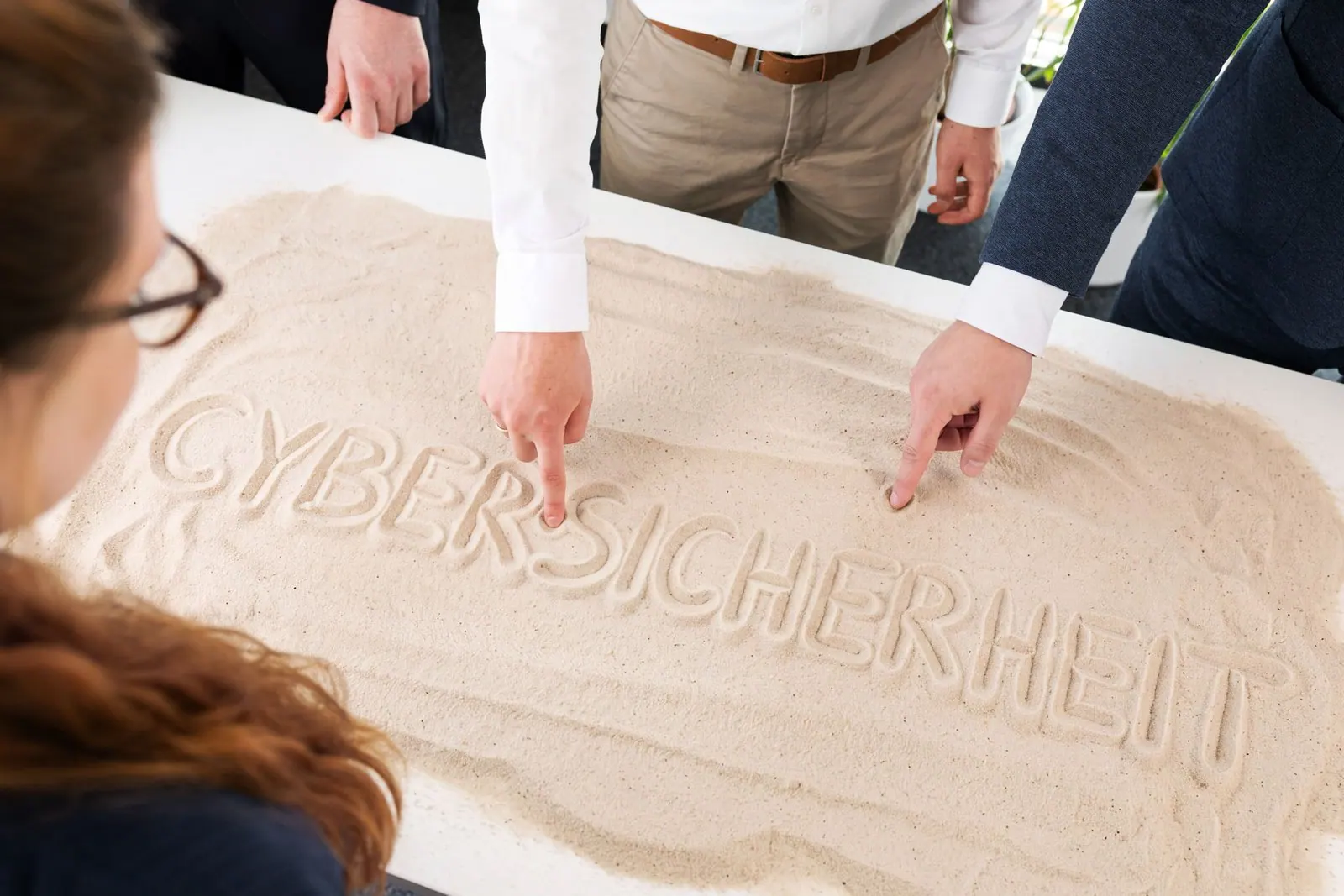 Mehrere Hände zeigen gemeinsam auf das in Sand geschriebene Wort CYBERSICHERHEIT