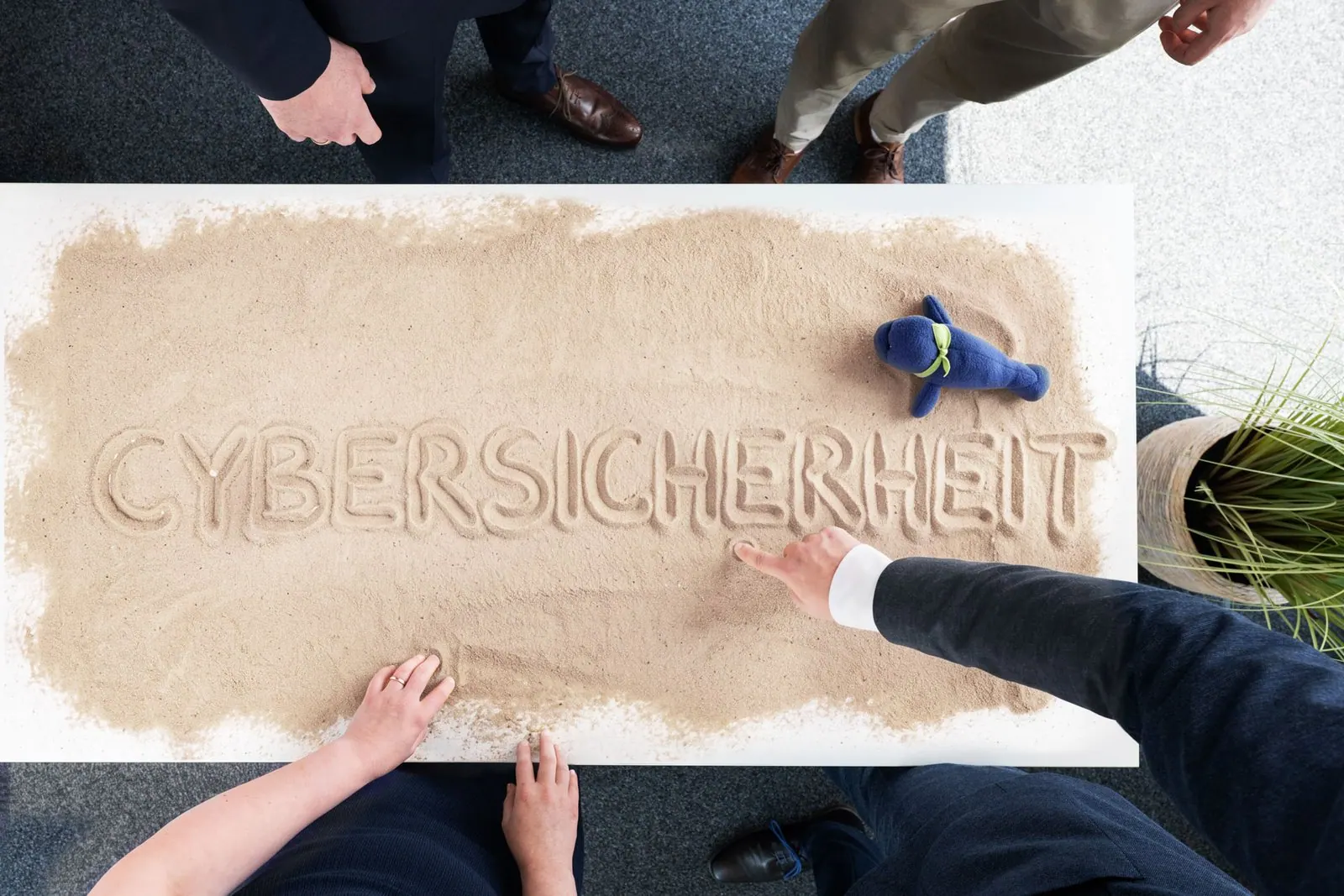 Draufsicht auf den Sandtisch mit dem Schriftzug CYBERSICHERHEIT, umgeben von mehreren Personen und der blauen Vera-Figur